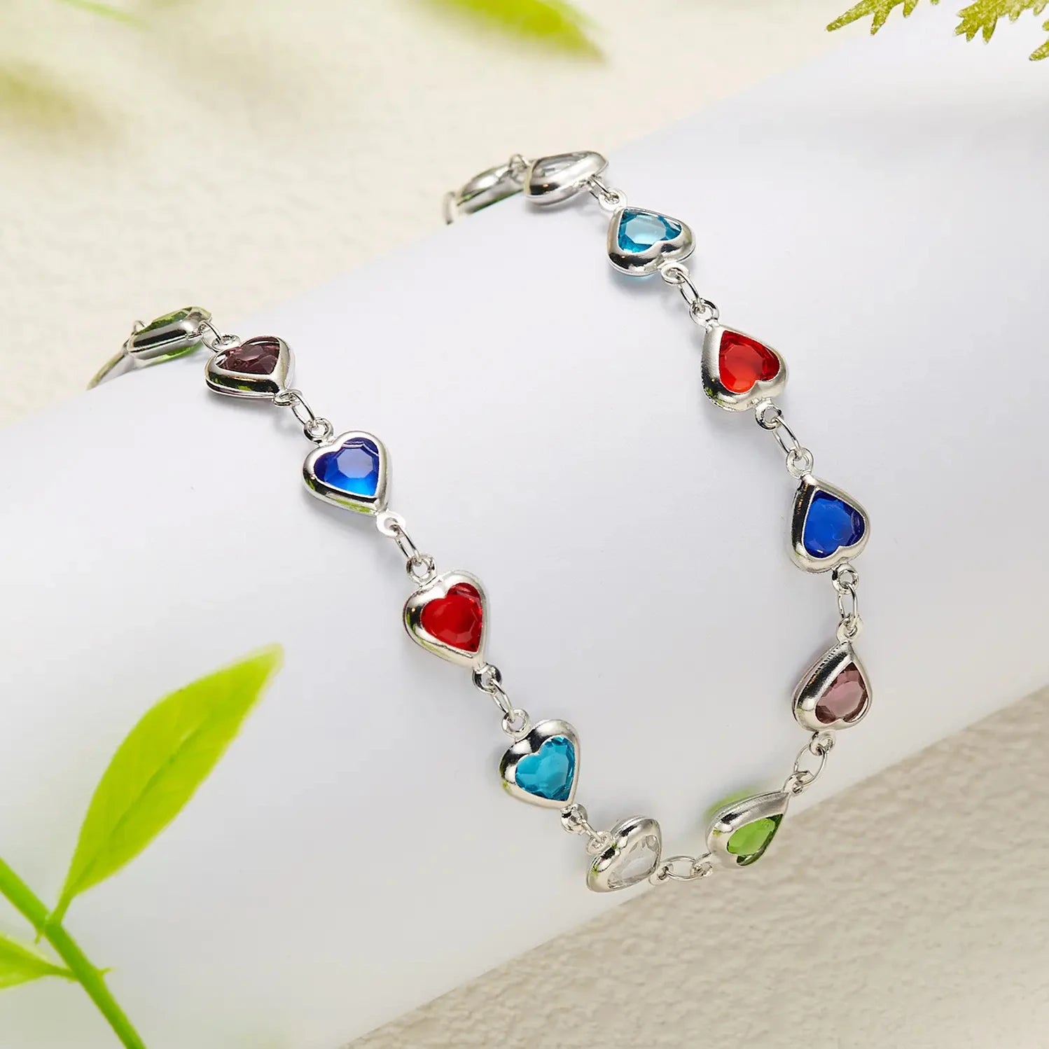Bracelet with heart-shaped colorful stones on a white surface with green leaves in the background